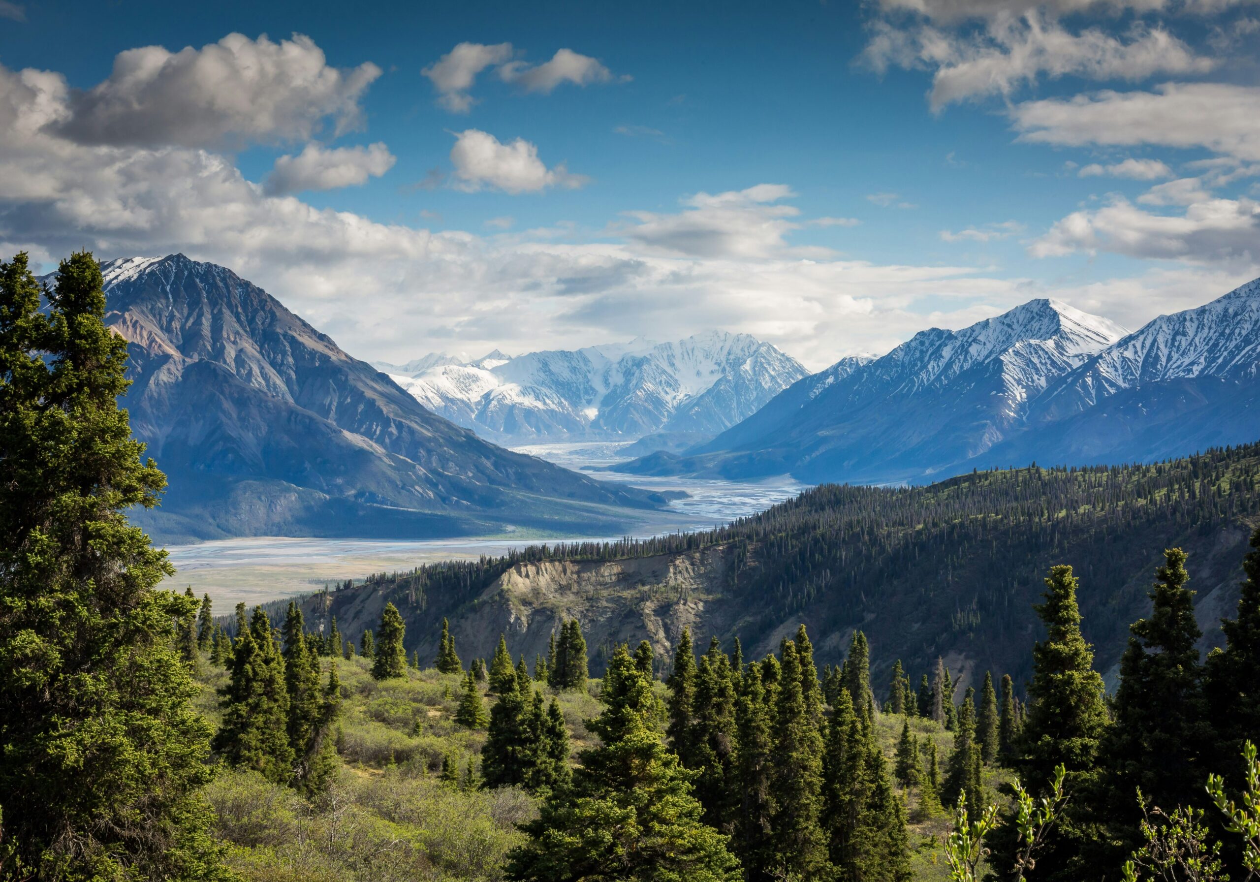 Mountain landscape with trees, snow-capped peaks, and cloudy sky.