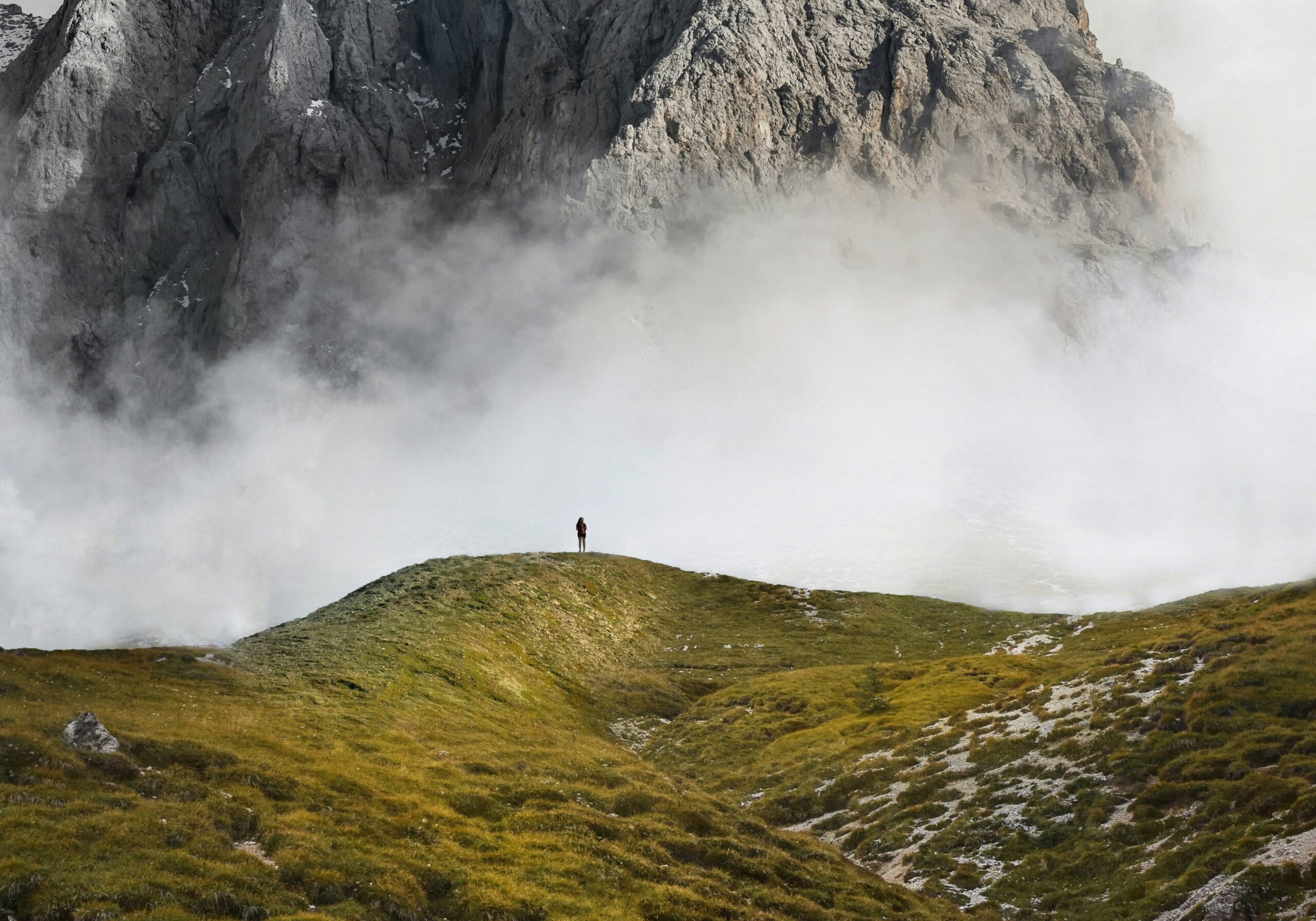 A person stands on a grassy hill below a misty, jagged mountain peak.