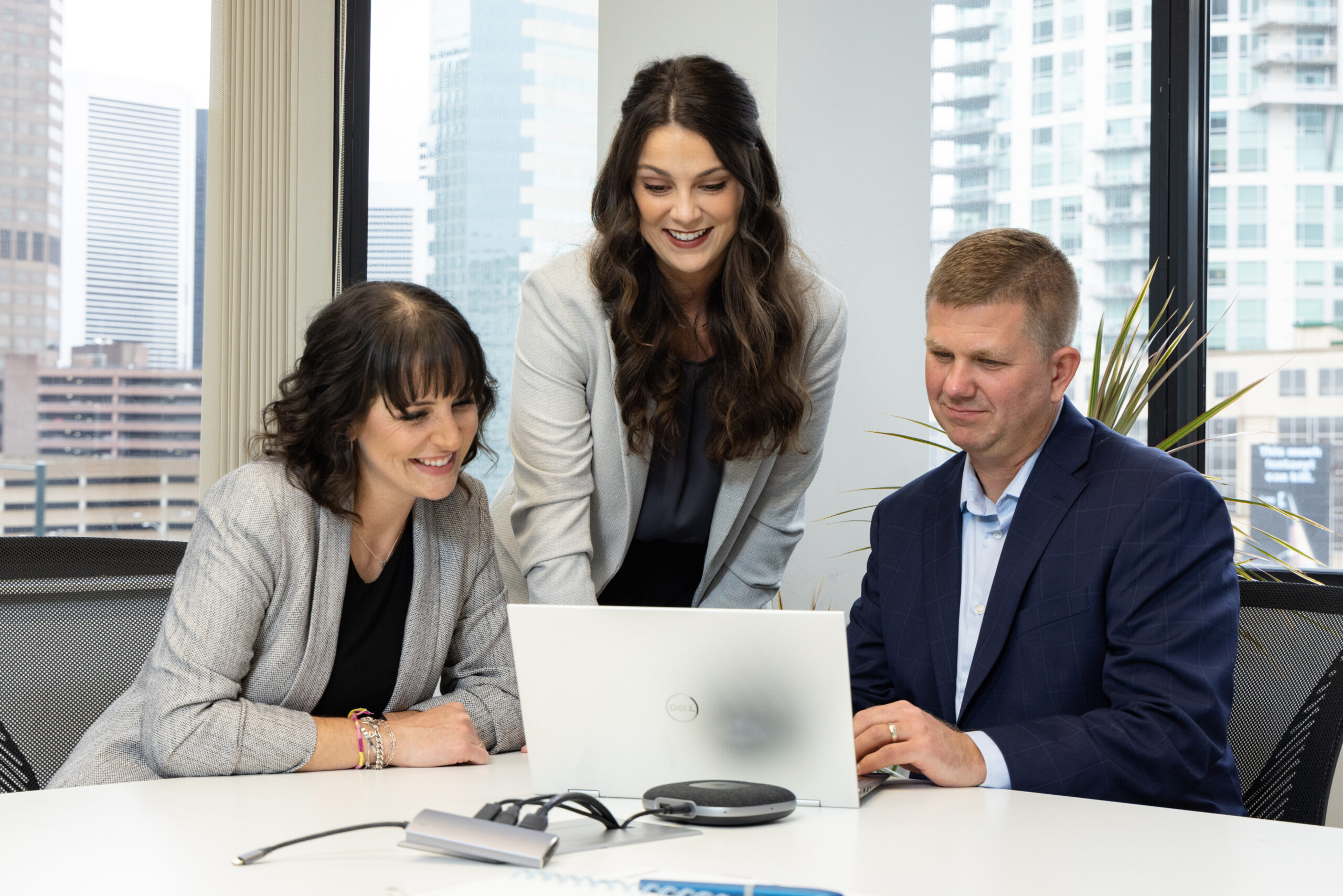 Three colleagues in business attire smiling and collaborating around a laptop in an office with cityscape windows
