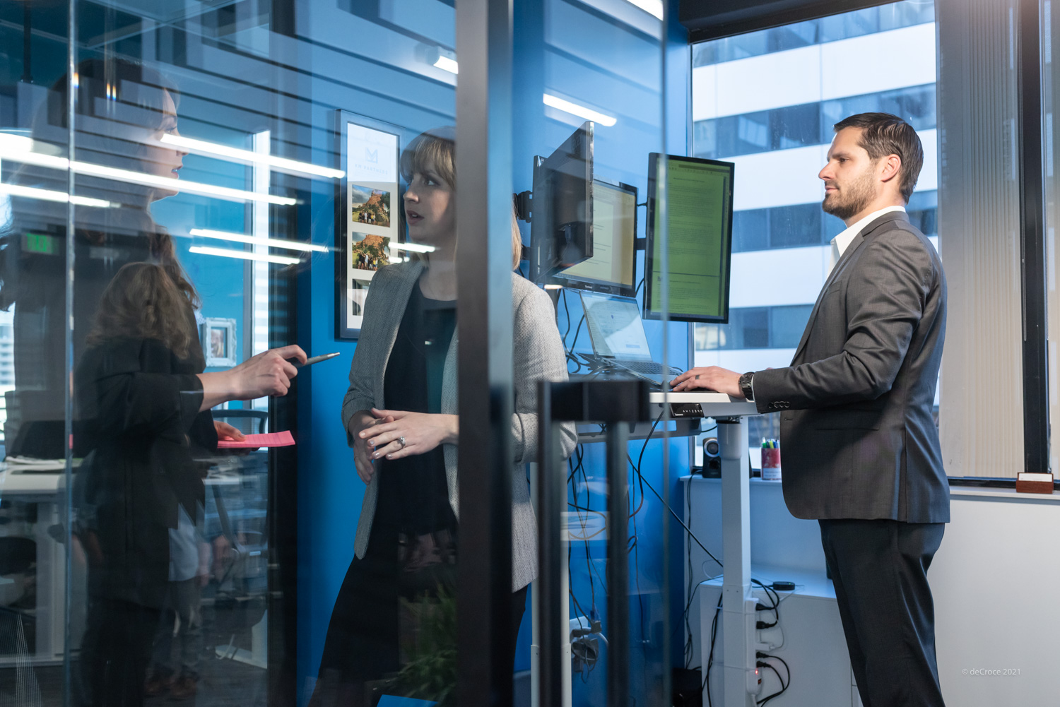 Two women discussing in a glass-walled office, one man working at a standing desk with dual monitors