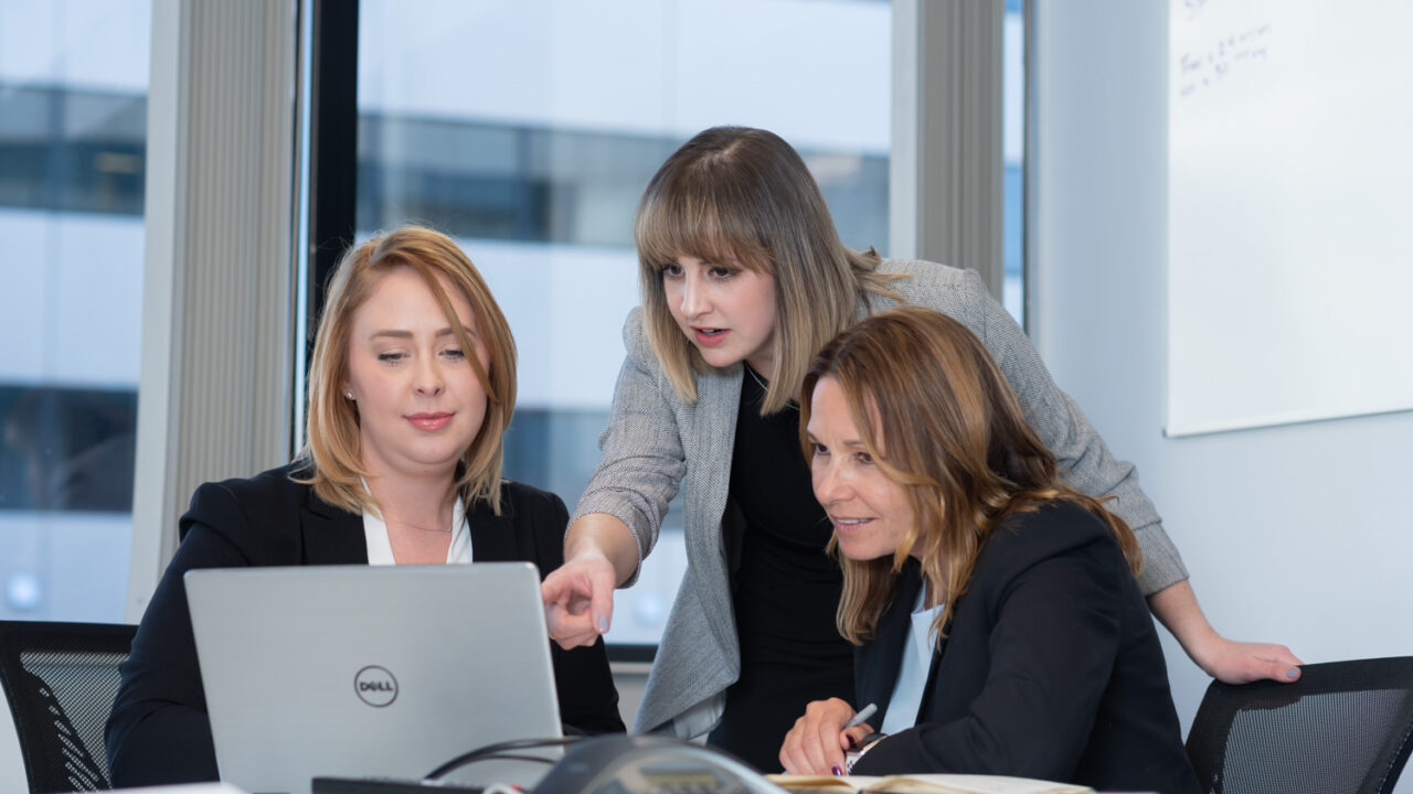 Three women collaborating at a conference table, one pointing at a laptop