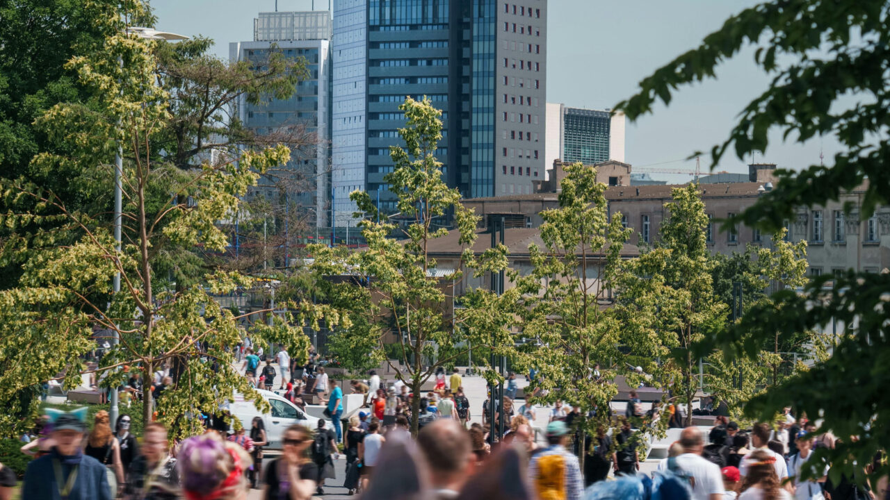 Crowded park with people walking, surrounded by trees, city skyscrapers in the background