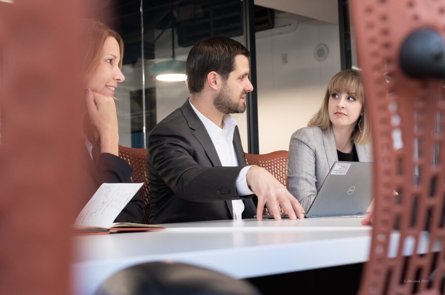 Three professionals in a meeting, one speaking, others listening, with laptop and notebook