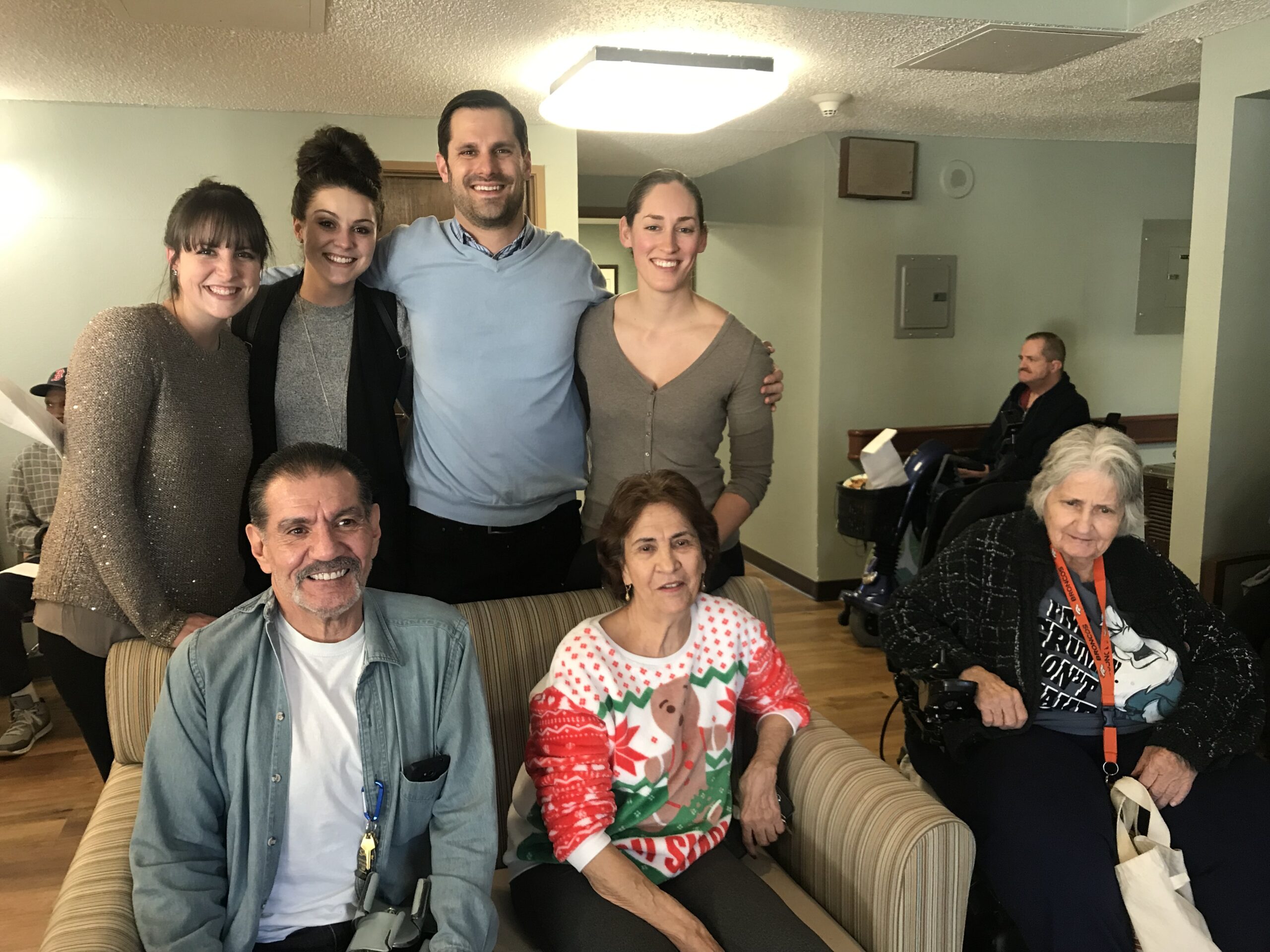 Group of seven people smiling in a cozy room, including Volunteering2.jpg individuals. Three are seated, and four stand closely behind