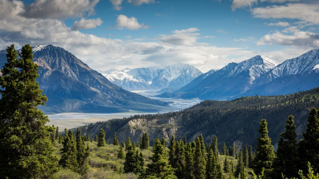Mountain landscape with trees, snow-capped peaks, and cloudy sky.