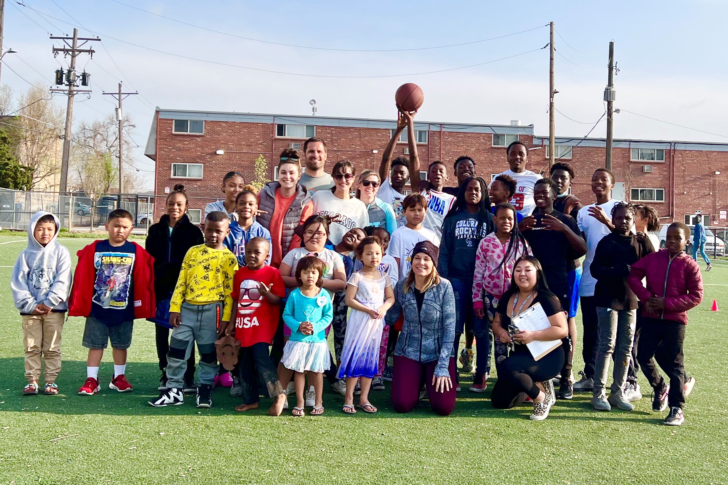 Children and adults gather outdoors, smiling as a child in the center holds up a basketball, suggesting teamwork and joy